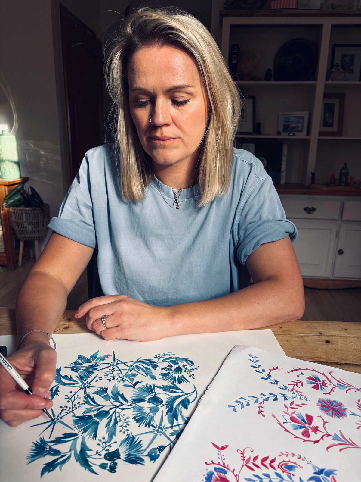 Anna Mcilwraith of To Pastures New sits at a wooden table, drawing intricate blue floral patterns on white paper with a black marker. Another sheet of paper with red and blue floral designs is visible in the foreground, set against a cozy home interior background.