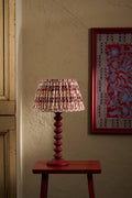 A table lamp with a red turned wood base and a pleated shade featuring a red, beige, and white geometric pattern, shown lit on a small red side table against a textured beige wall.