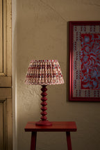 A table lamp with a red turned wood base and a pleated shade featuring a red, beige, and white geometric pattern, shown lit on a small red side table against a textured beige wall.