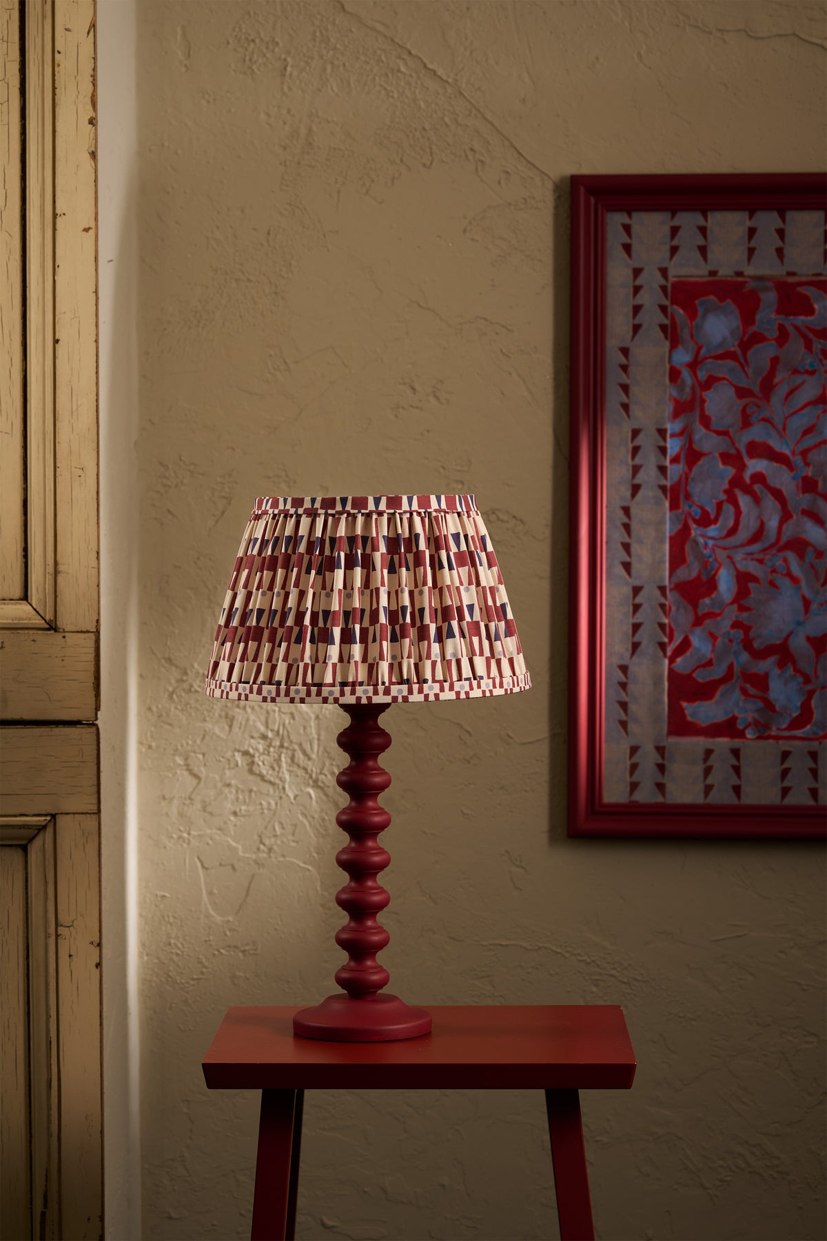A table lamp with a red turned wood base and a pleated shade featuring a red, beige, and white geometric pattern, shown lit on a small red side table against a textured beige wall.