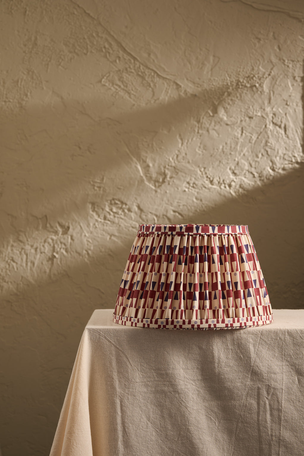A pleated fabric lampshade with a geometric pattern of red, blue, and cream stripes sits on a draped beige tablecloth against a textured beige plaster wall.
