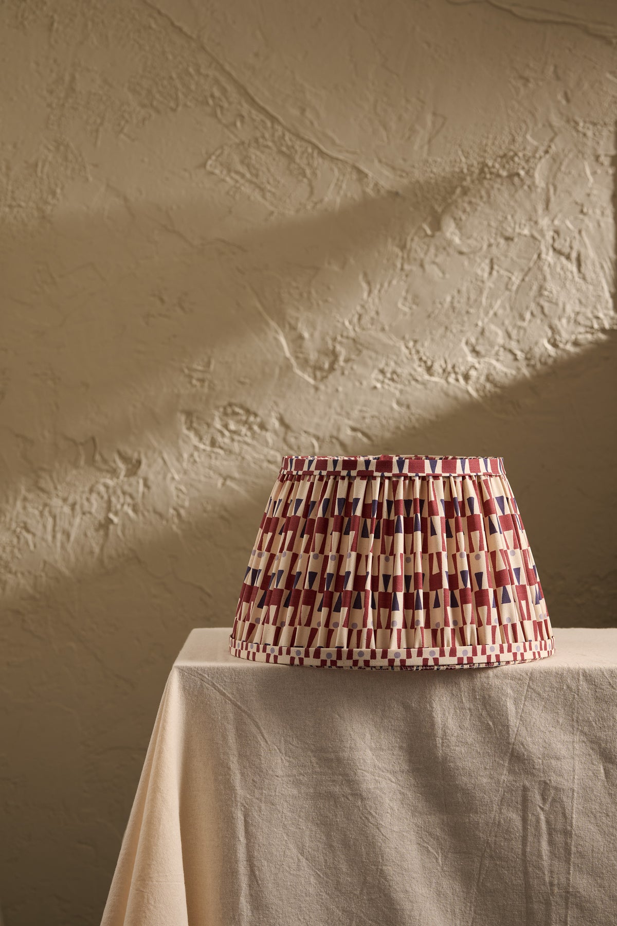 A pleated fabric lampshade with a geometric pattern of red, blue, and cream stripes sits on a draped beige tablecloth against a textured beige plaster wall.