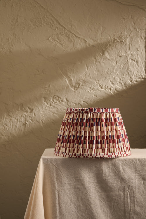 A pleated fabric lampshade with a geometric pattern of red, blue, and cream stripes sits on a draped beige tablecloth against a textured beige plaster wall.