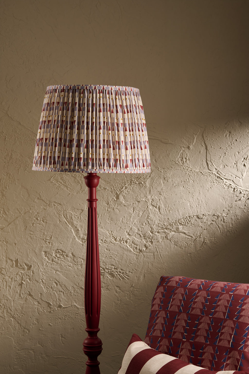 A tall floor lamp with a pleated fabric shade in a light blue, red, and white pattern, mounted on a dark red wooden stand, set against a textured beige wall with a red patterned armchair partially visible.