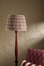 A tall floor lamp with a pleated fabric shade in a light blue, red, and white pattern, mounted on a dark red wooden stand, set against a textured beige wall with a red patterned armchair partially visible.