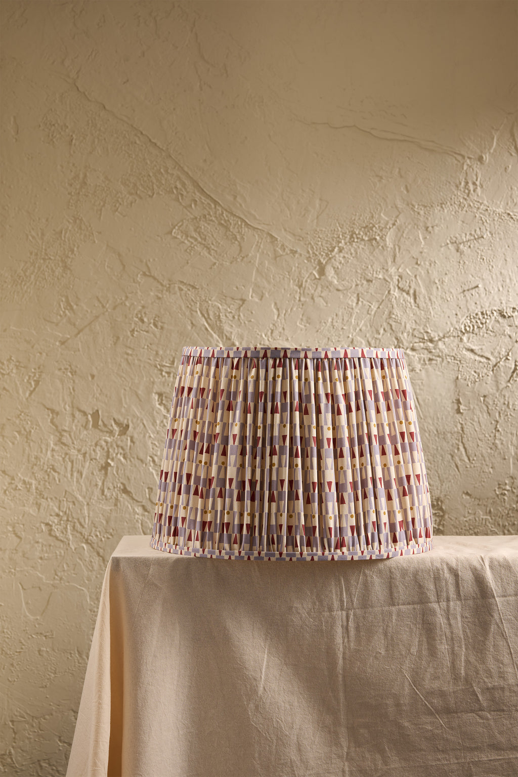A pleated fabric lampshade with a multicolor geometric pattern in light blue, and red, displayed on a draped table against a textured beige wall.