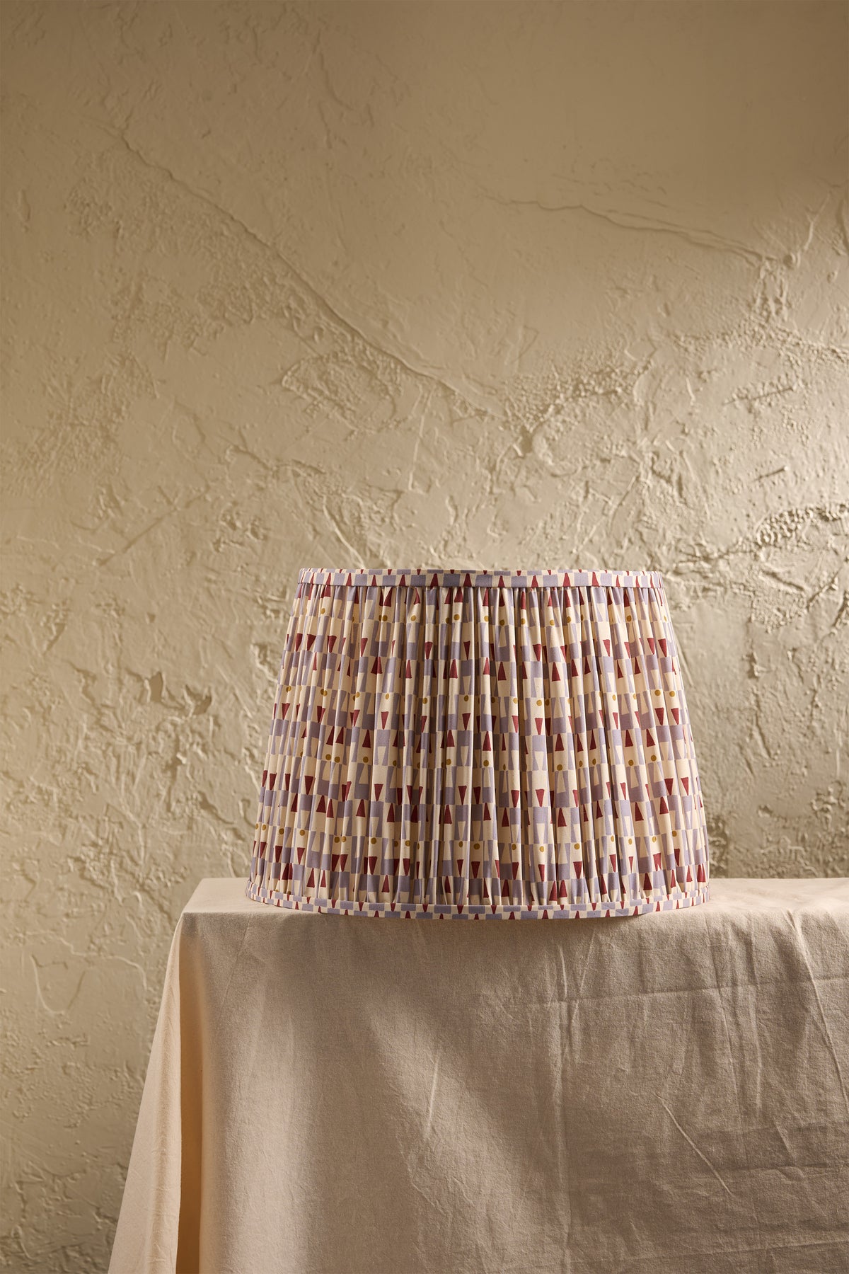 A pleated fabric lampshade with a multicolor geometric pattern in light blue, and red, displayed on a draped table against a textured beige wall.