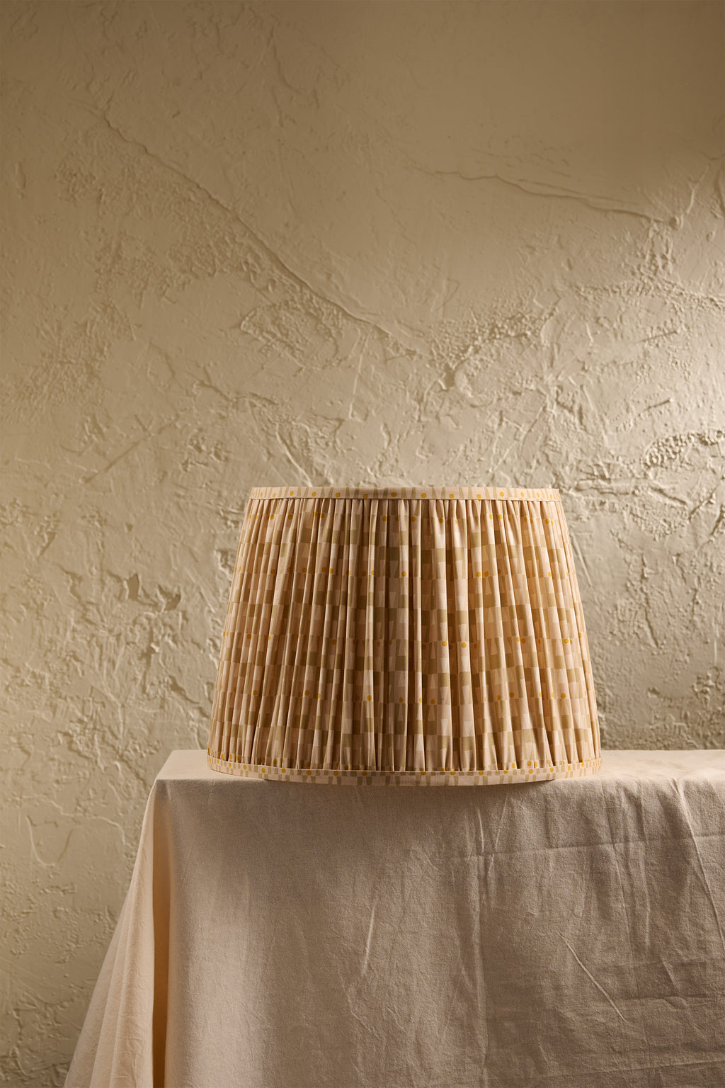 A pleated lampshade in an oat and ochre pattern, displayed on a draped table against a textured beige plaster wall.
