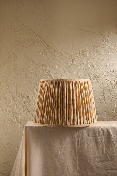 A pleated lampshade in an oat and ochre pattern, displayed on a draped table against a textured beige plaster wall.