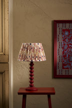 A table lamp with a to pastures new pleated fabric shade featuring a red, ochre, and blue geometric pattern, mounted on a dark red turned wood pedestal base. The lamp is styled on a matching dark red side table against a textured beige wall, with a framed artwork partially visible on the right.