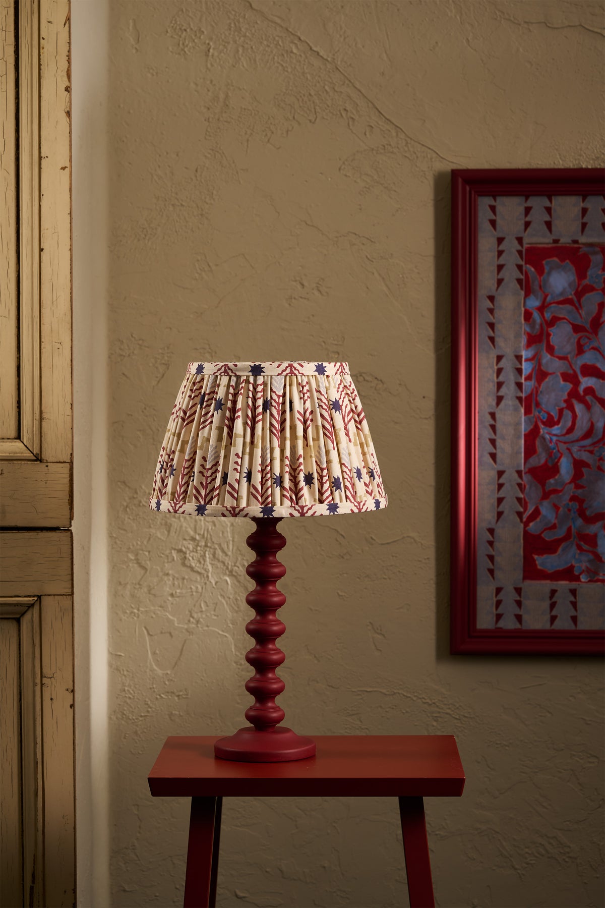 A table lamp with a to pastures new pleated fabric shade featuring a red, ochre, and blue geometric pattern, mounted on a dark red turned wood pedestal base. The lamp is styled on a matching dark red side table against a textured beige wall, with a framed artwork partially visible on the right.