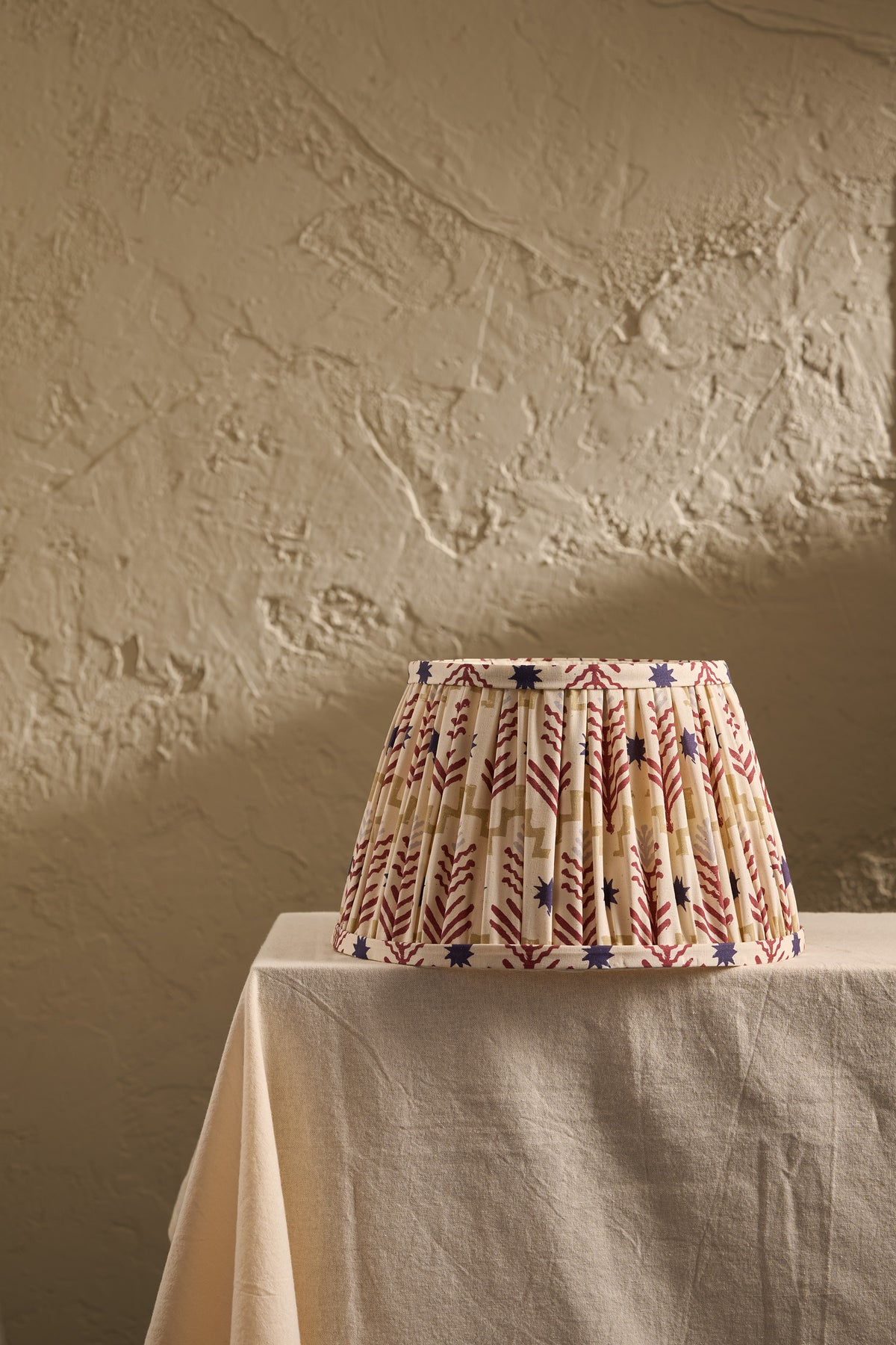A pleated fabric lampshade with a cream base and a red, blue, and ochre geometric pattern, displayed on a draped table against a textured beige plaster wall.