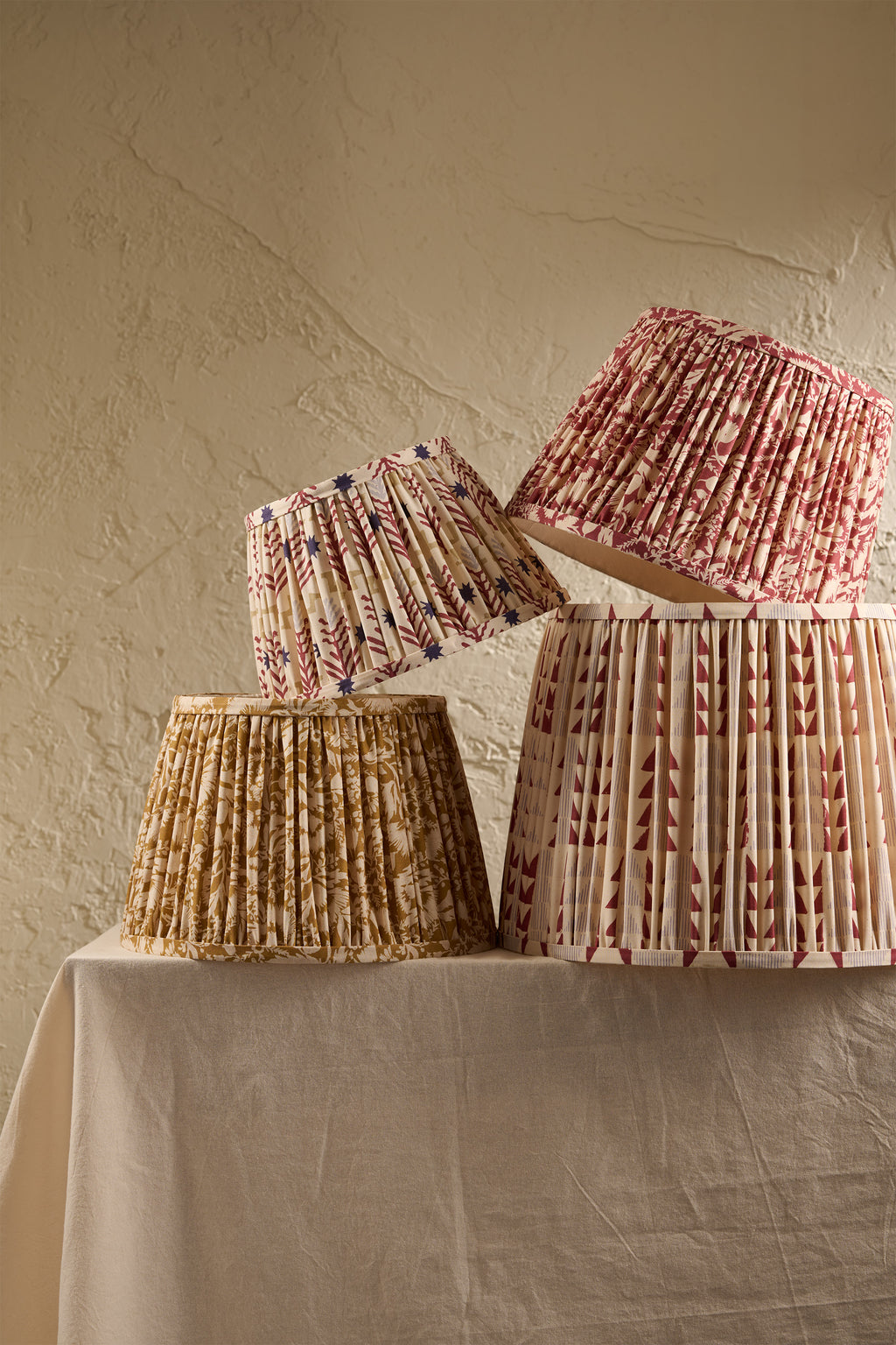 Four pleated fabric lampshades in different floral and geometric patterns, displayed on a draped cream tablecloth against a textured beige plaster wall.