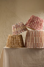 Four pleated fabric lampshades in different floral and geometric patterns, displayed on a draped cream tablecloth against a textured beige plaster wall.