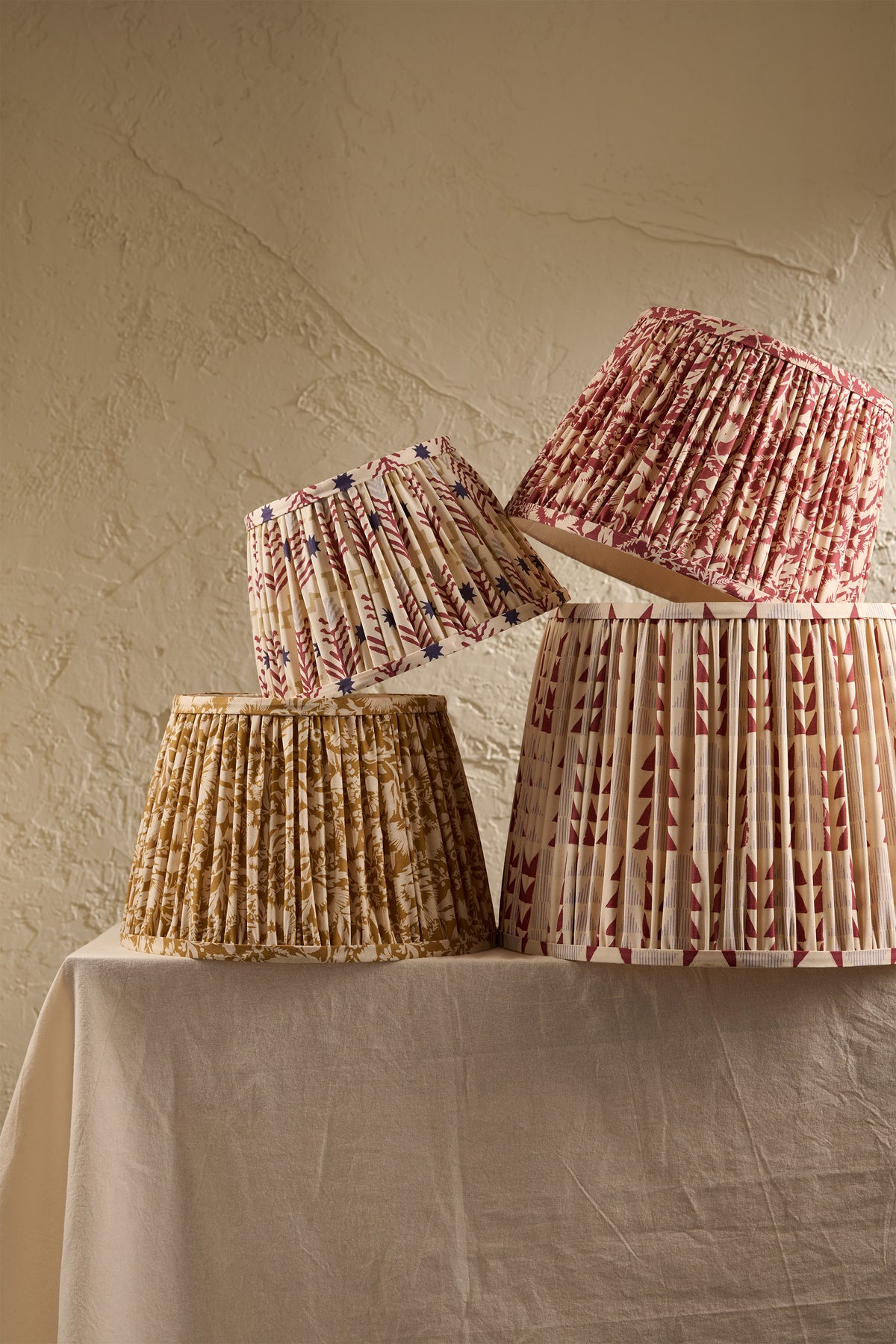 Four pleated fabric lampshades in different floral and geometric patterns, displayed on a draped cream tablecloth against a textured beige plaster wall.