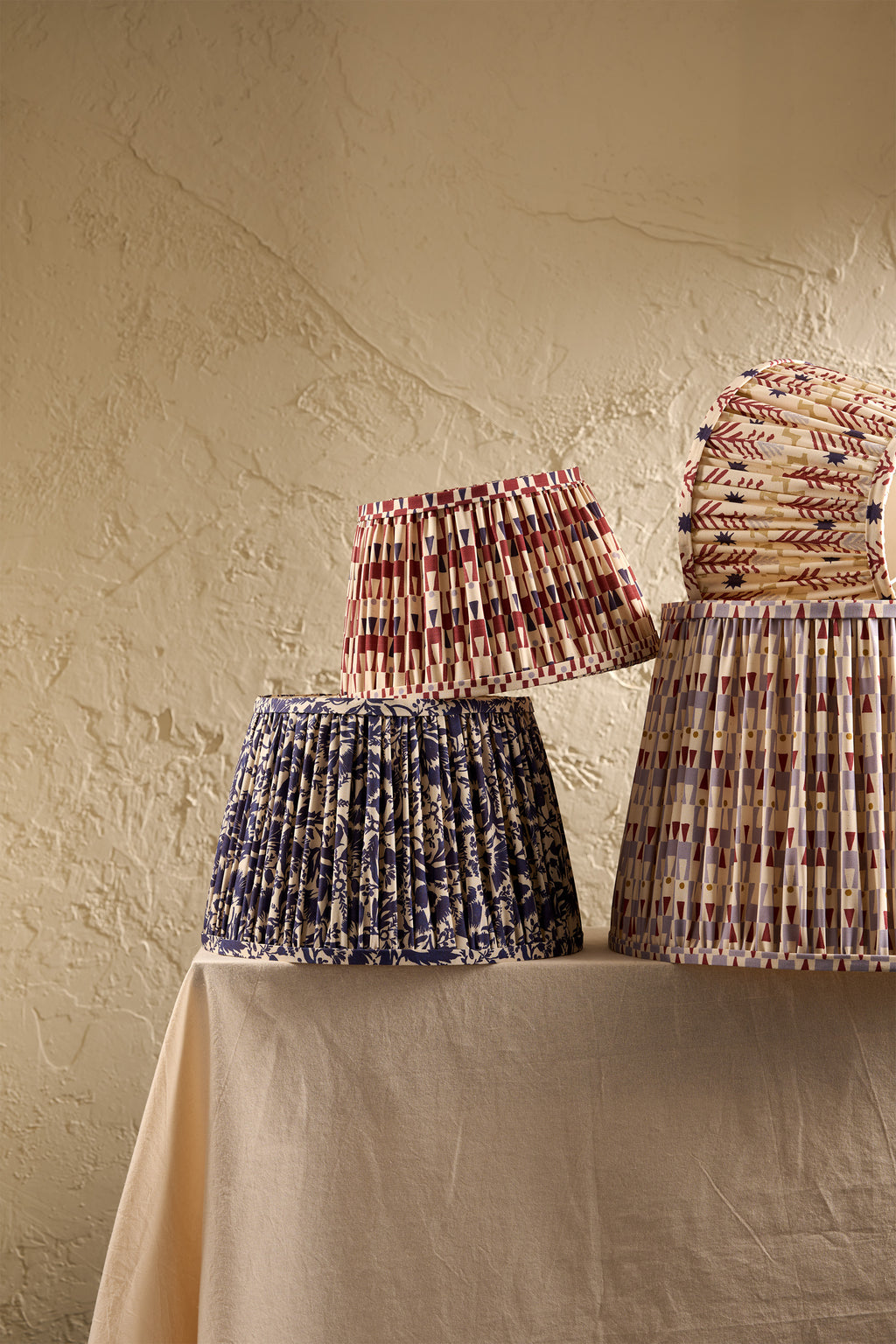 Three pleated fabric lampshades in different patterns and colors displayed on a draped table against a textured beige wall.