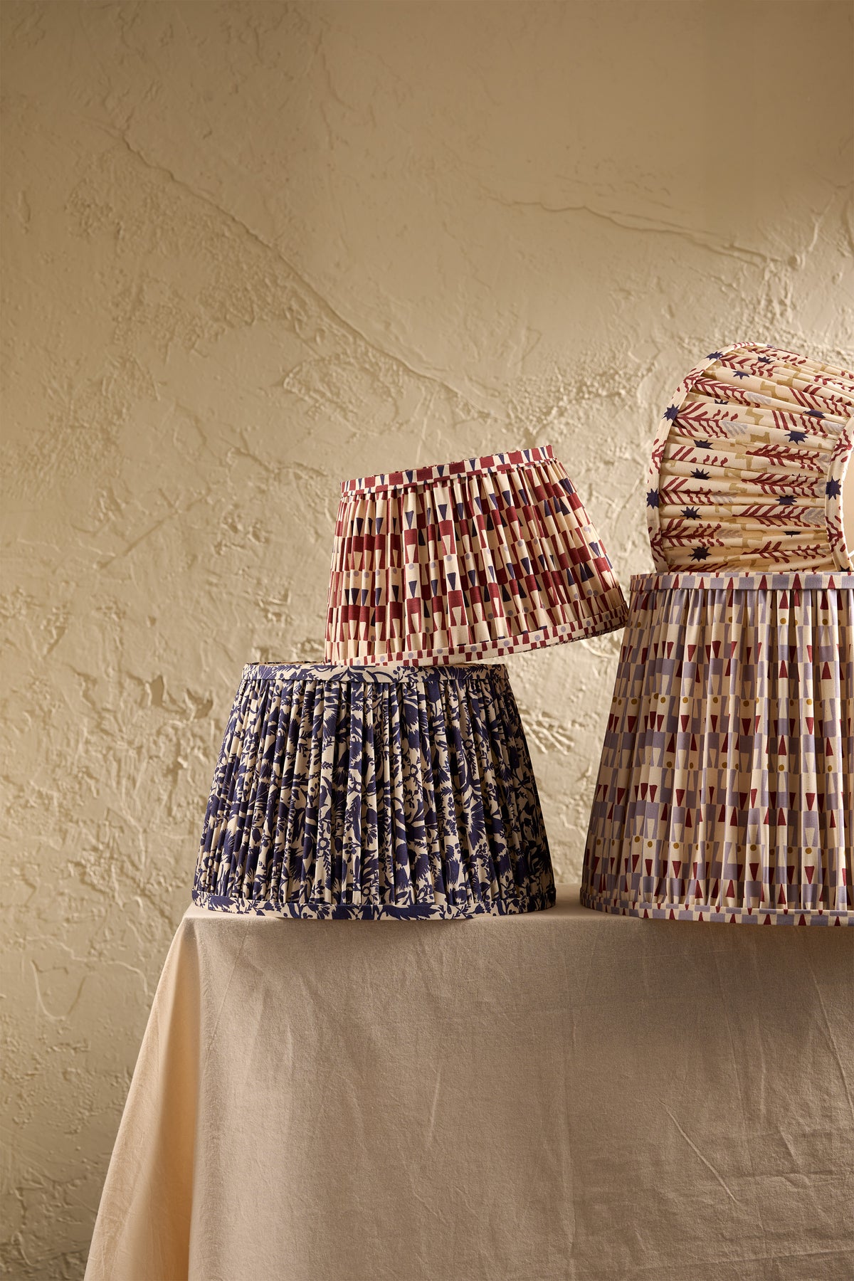 Three pleated fabric lampshades in different patterns and colors displayed on a draped table against a textured beige wall.