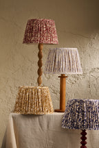 Three decorative table lamps with pleated fabric shades in different colors and patterns, displayed on a draped table against a textured beige wall.