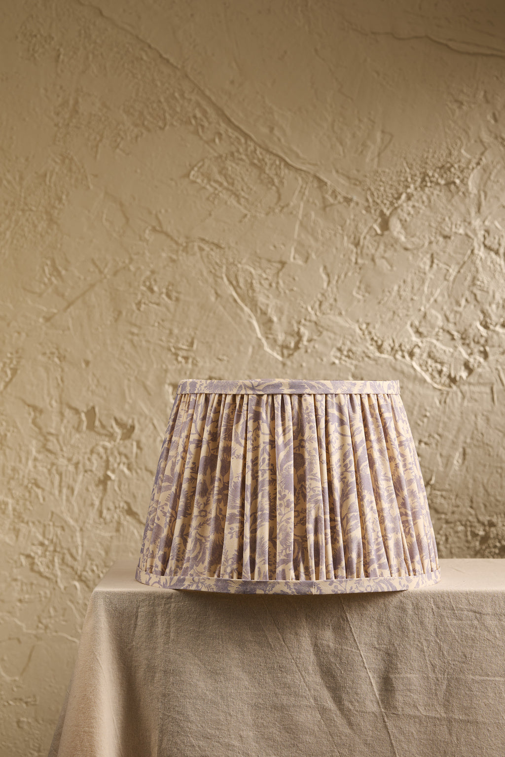 A pleated fabric lampshade with a subtle beige and lavender floral pattern, displayed on a draped table against a textured beige plaster wall.