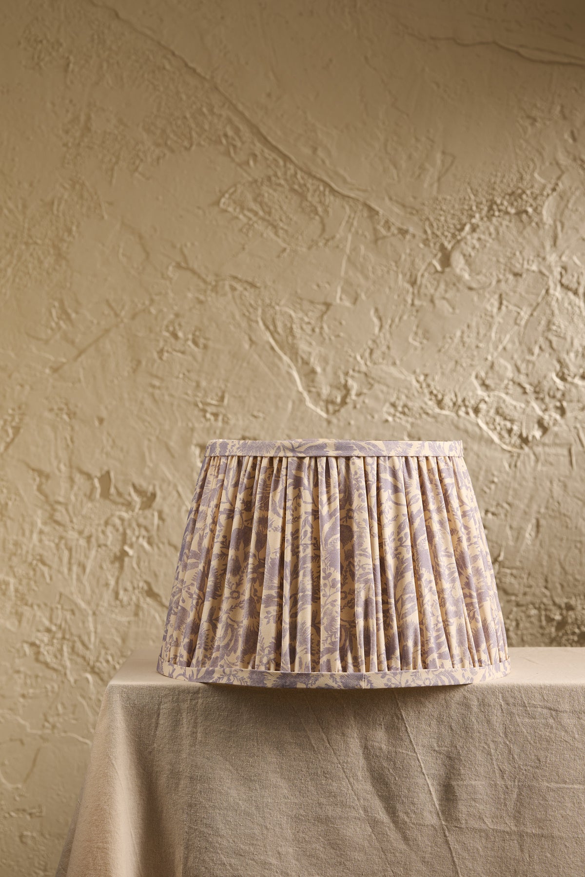 A pleated fabric lampshade with a subtle beige and lavender floral pattern, displayed on a draped table against a textured beige plaster wall.