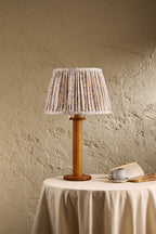 A table lamp with a pleated fabric shade in a beige and white pattern and a round wooden base, displayed on a draped table against a textured beige wall.