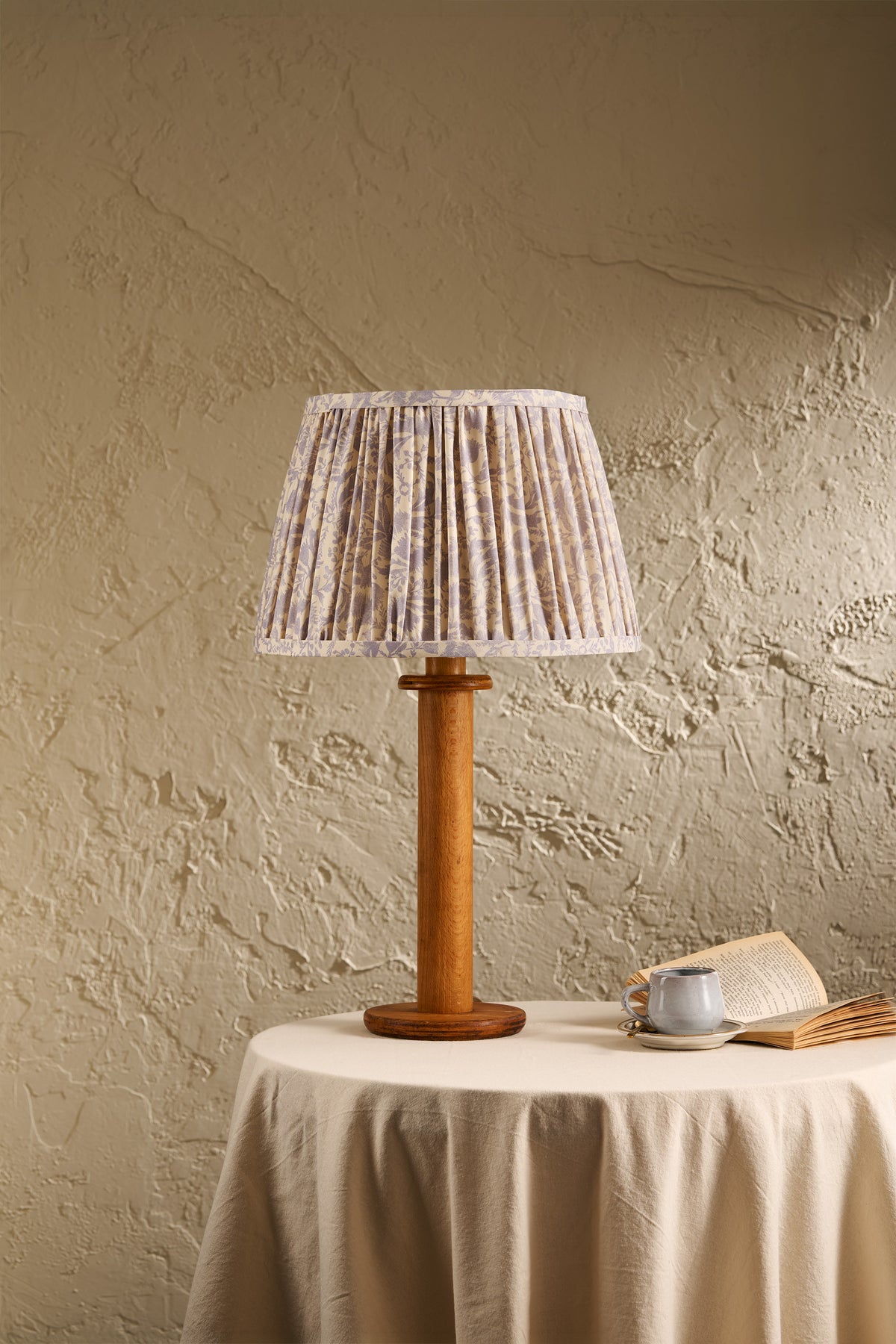 A table lamp with a pleated fabric shade in a beige and white pattern and a round wooden base, displayed on a draped table against a textured beige wall.