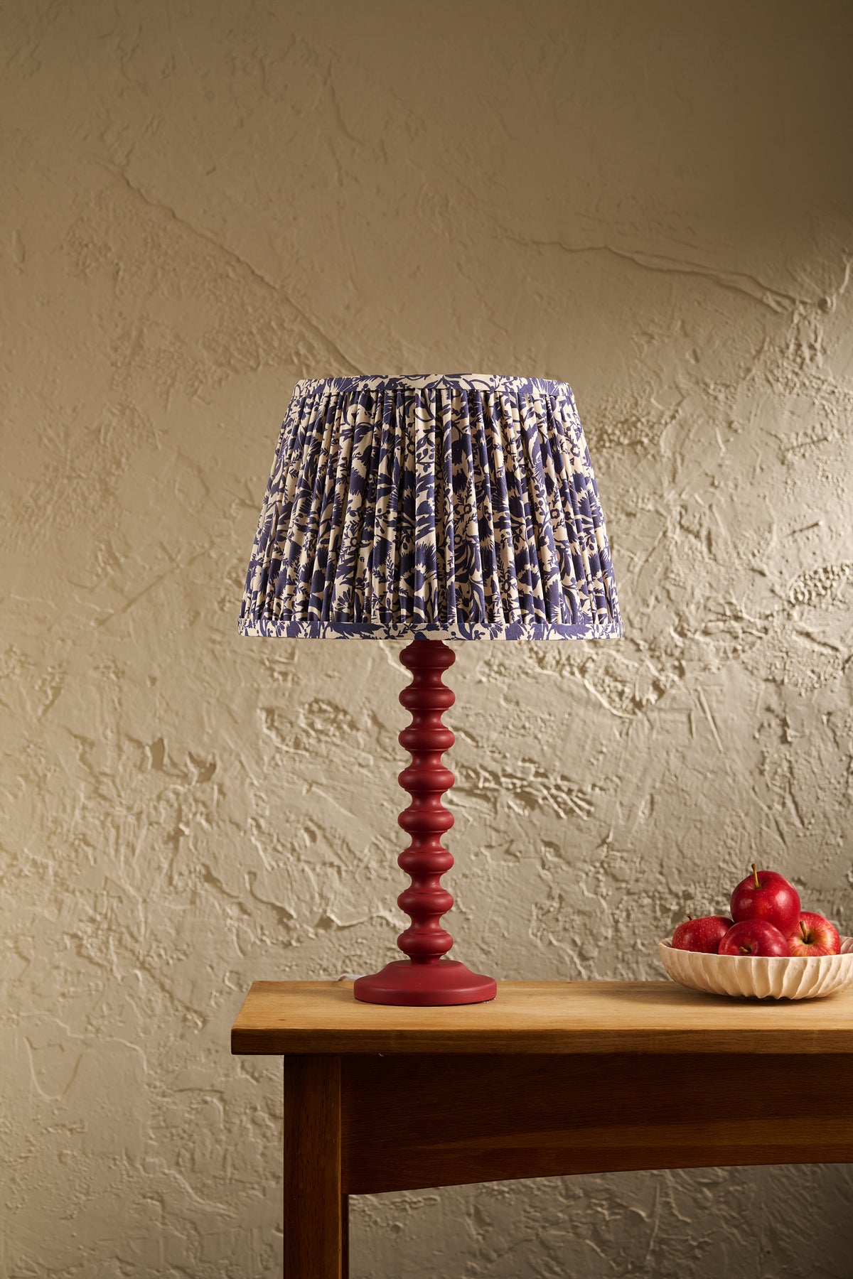 A table lamp with a pleated blue and white patterned fabric shade and a dark red spiral pedestal base, set on a wooden table against a textured beige wall with a bowl of red apples nearby.