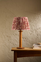 A table lamp with a pleated pink and red floral fabric shade and a natural wood base, set on a wooden table against a textured beige wall.