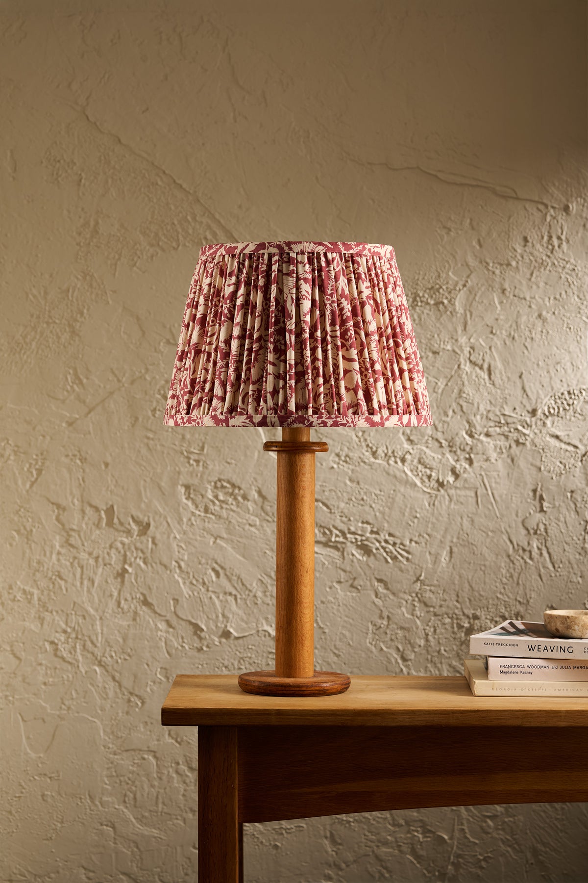 A table lamp with a pleated pink and red floral fabric shade and a natural wood base, set on a wooden table against a textured beige wall.