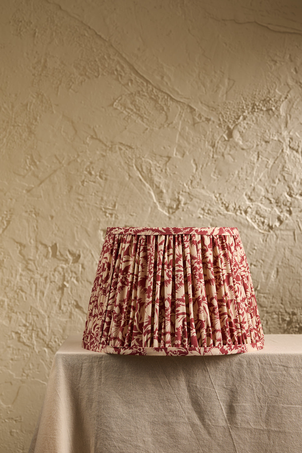 A pleated fabric lampshade with a red and beige floral toile pattern, displayed on a draped table against a textured beige plaster wall.