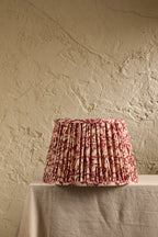 A pleated fabric lampshade with a red and beige floral toile pattern, displayed on a draped table against a textured beige plaster wall.