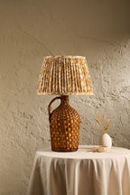 A rustic table lamp with a woven brown handle and a pleated beige and brown patterned shade, displayed on a draped white tablecloth against a textured beige wall.