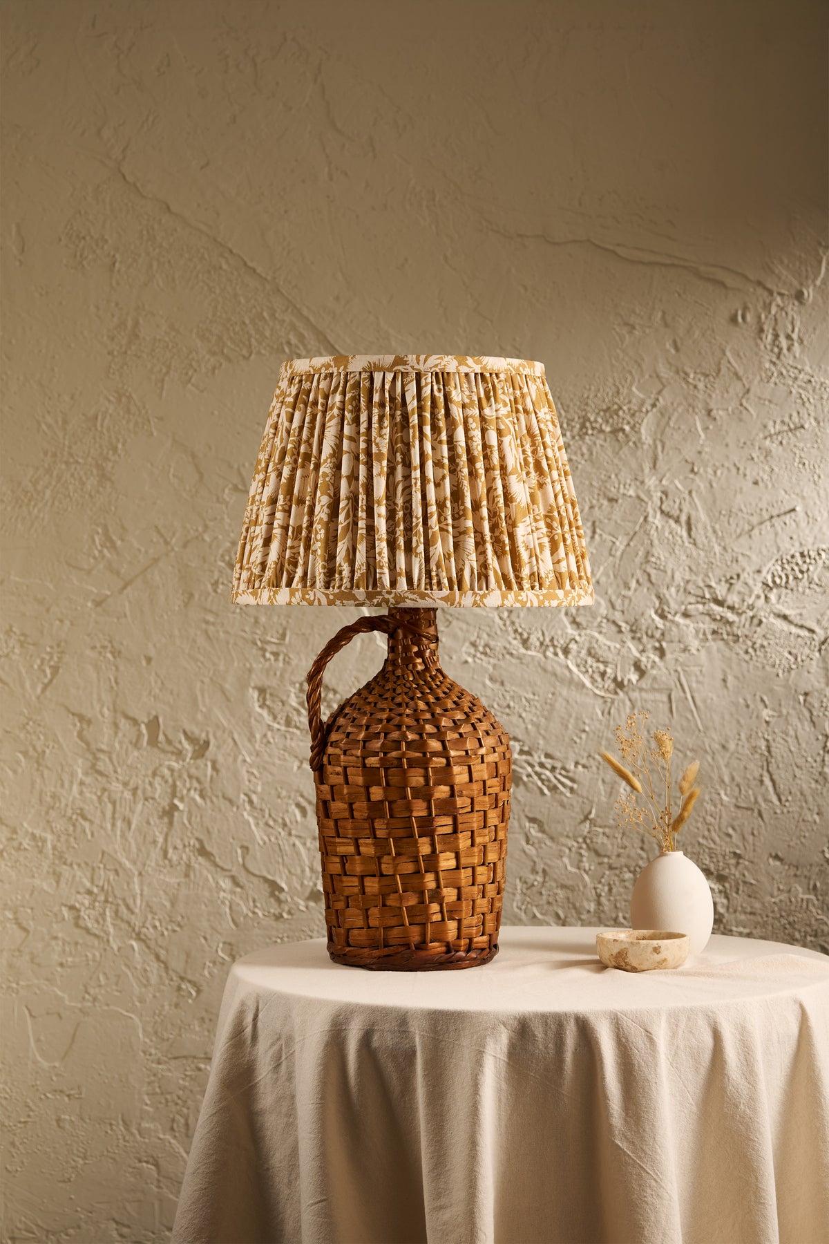 A rustic table lamp with a woven brown handle and a pleated beige and brown patterned shade, displayed on a draped white tablecloth against a textured beige wall.