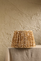 A pleated fabric lampshade with a warm brown and beige floral pattern, displayed on a draped table against a textured beige plaster wall.