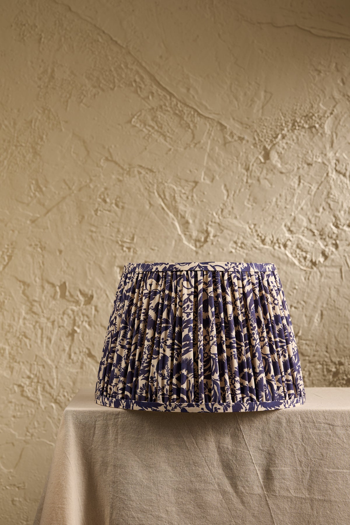 A pleated fabric lampshade with a blue and beige floral pattern, displayed on a draped table against a textured beige plaster wall.