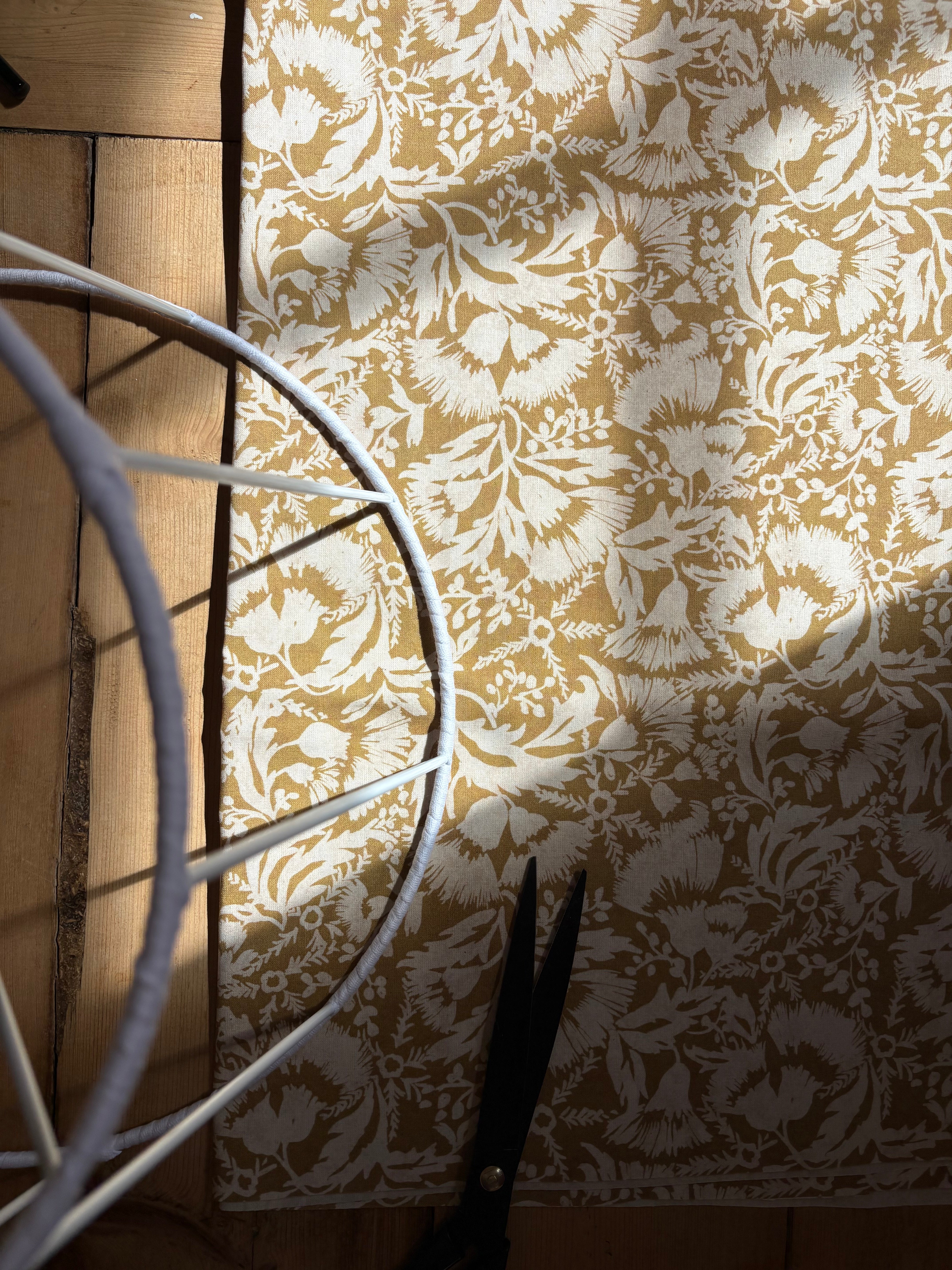 A close-up of a beige and white damask-patterned fabric or rug laid on a wooden floor, with a white metal circular frame partially visible on the left and black-handled scissors at the bottom right.
